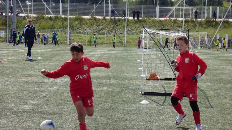 Deux jeunes joueurs en maillot rouge s'entraînent au football sur un terrain en herbe synthétique, avec d'autres enfants et un entraîneur en arrière-plan.