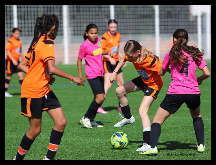 Des jeunes joueuses de football en maillots oranges et roses disputent le ballon sur un terrain en gazon synthétique.