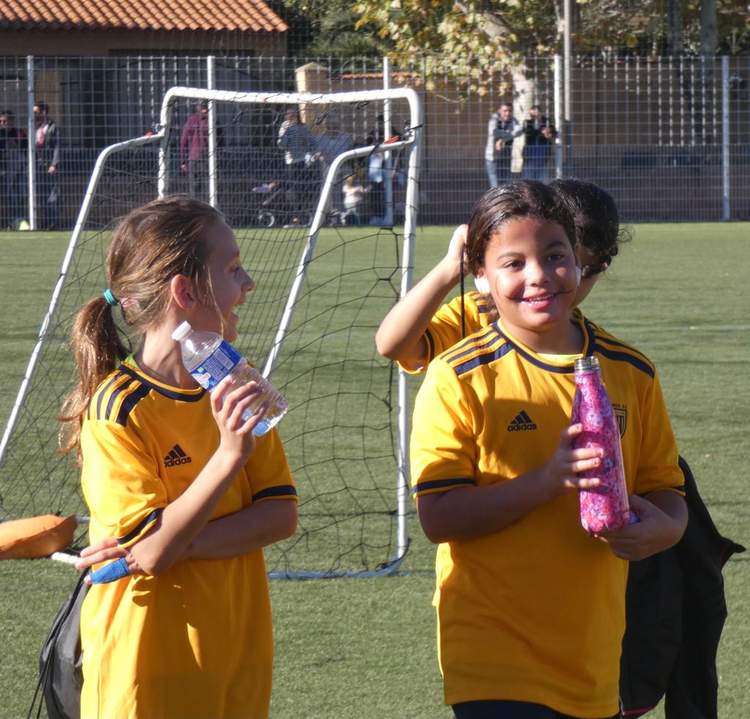 Deux jeunes joueuses en maillot jaune se désaltèrent sur le terrain de football, avec un but en arrière-plan.