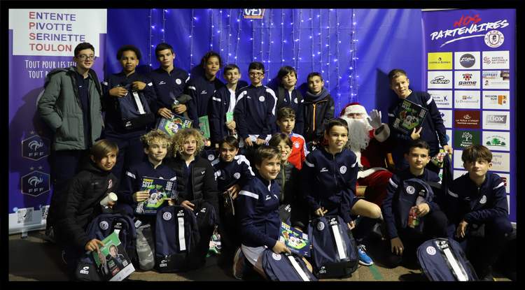 Un groupe de jeunes footballeurs pose avec des cadeaux et le Père Noël devant un mur de partenaires.