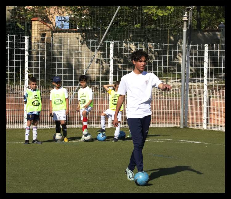Un jeune entraîneur guide des enfants lors d'une séance de football sur un terrain synthétique.