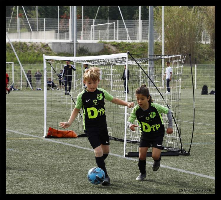Deux jeunes joueurs de football en maillot vert et noir sont en pleine action sur un terrain, avec un ballon bleu à leurs pieds et des buts en arrière-plan.