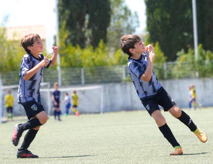 Deux jeunes joueurs de football en maillot gris et noir sont en pleine action sur un terrain vert, avec d'autres joueurs flous en arrière-plan.