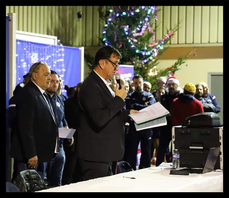 Un homme en costume parle au micro devant une assemblée lors d'un événement festif avec un sapin de Noël en arrière-plan.