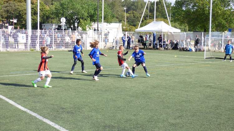 Des jeunes joueurs de football en pleine action sur un terrain synthétique, avec des spectateurs en arrière-plan.