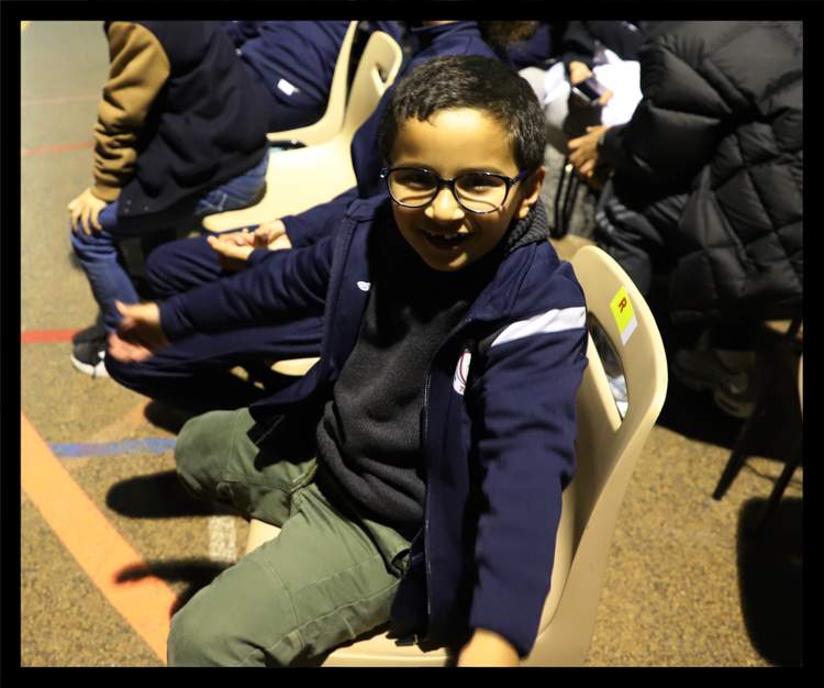 Un jeune garçon souriant avec des lunettes est assis sur une chaise, entouré d'autres personnes.