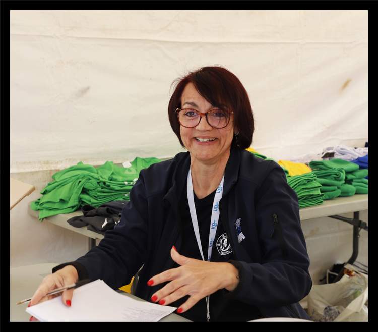 Une femme souriante avec des lunettes est assise à une table, tenant un stylo et des papiers, avec des piles de t-shirts colorés en arrière-plan.