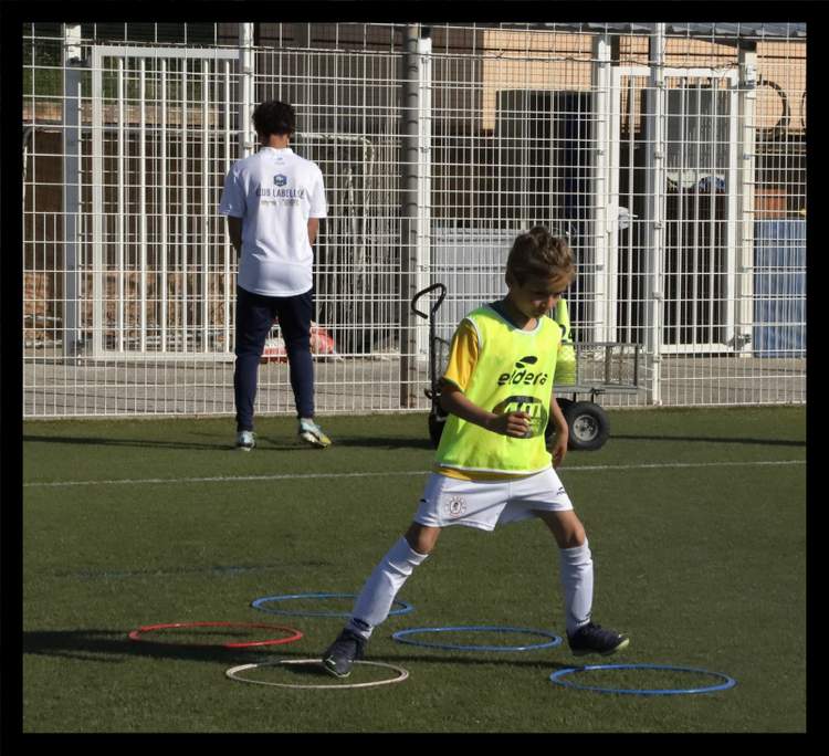 Un jeune joueur de football s'entraîne sur le terrain avec des cerceaux, tandis qu'un autre joueur se tient en arrière-plan.