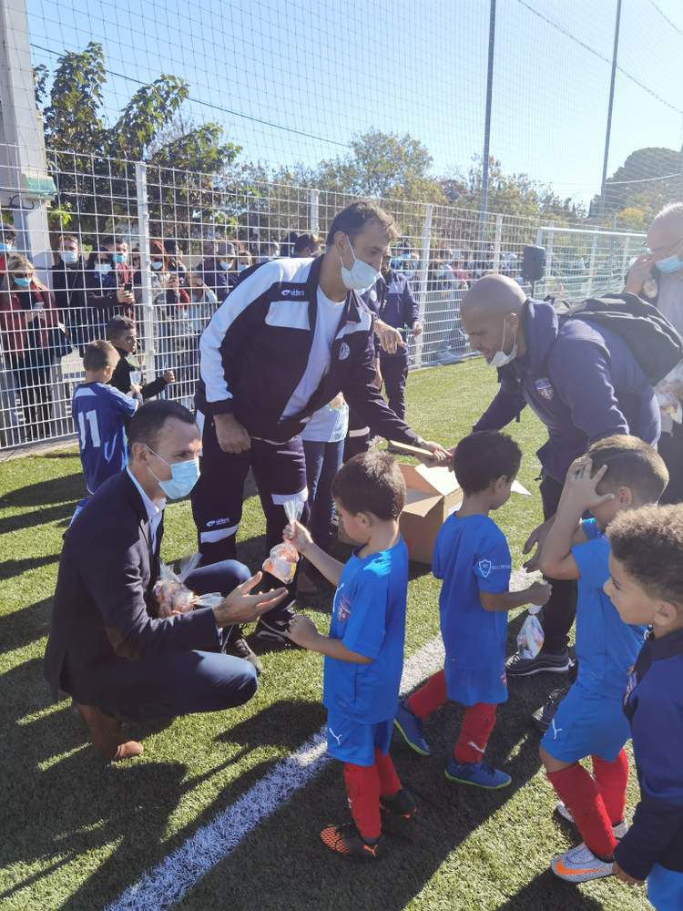 Des adultes masqués distribuent des cadeaux à de jeunes footballeurs sur un terrain ensoleillé, avec du public en arrière-plan.