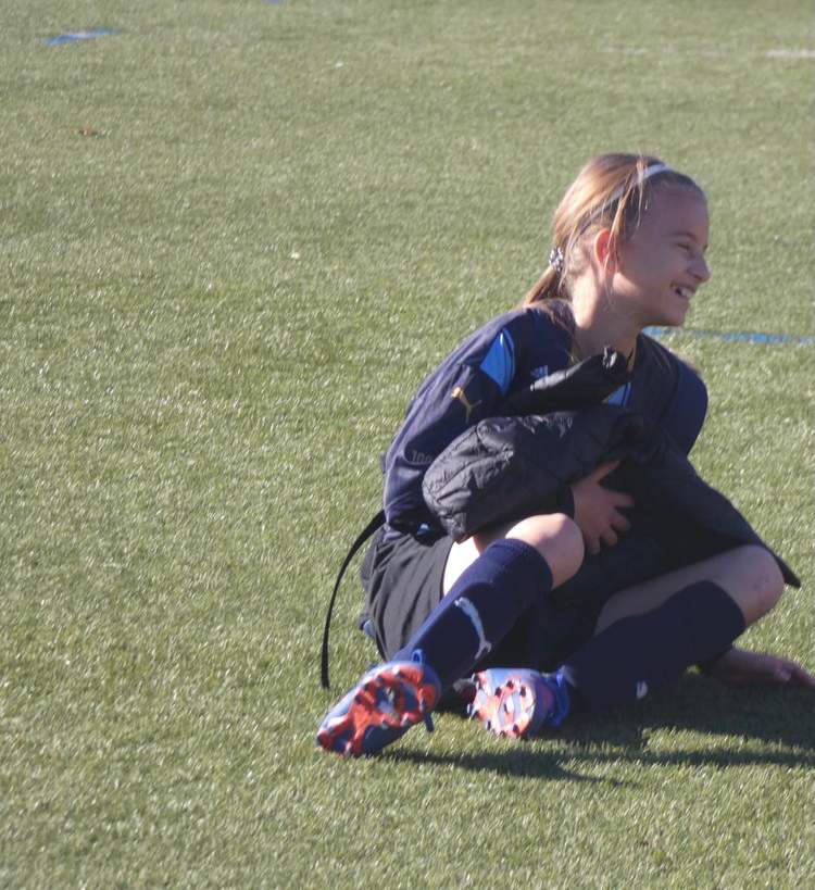 Une jeune joueuse de football est assise sur le terrain, souriante et tenant une veste.