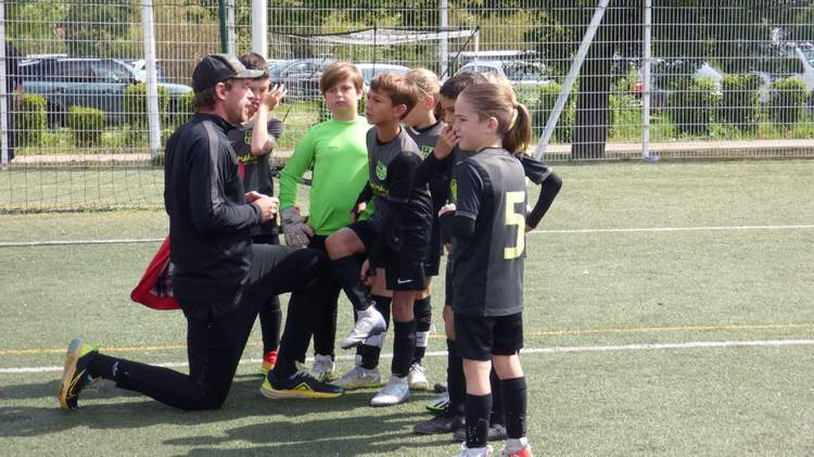 Un entraîneur à genoux parle à un groupe de jeunes joueurs de football sur un terrain en gazon synthétique.