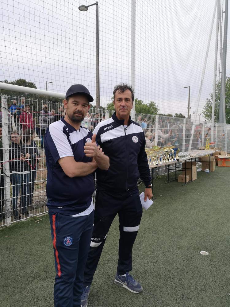 Deux hommes posent sur un terrain de football, avec des trophées en arrière-plan et des spectateurs derrière une clôture.