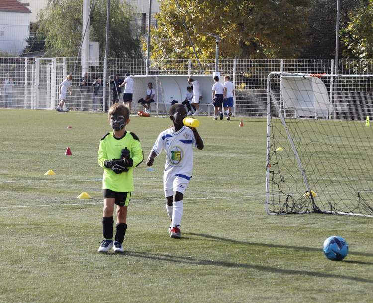 Deux jeunes joueurs de football marchent sur le terrain, l'un buvant à la bouteille, avec d'autres joueurs en arrière-plan.