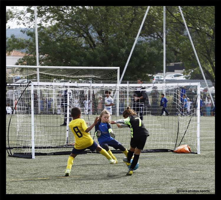 Trois jeunes joueurs de football en pleine action devant un but, avec d'autres personnes en arrière-plan.