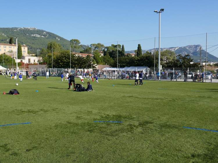 Des jeunes joueurs de football s'entraînent sur un terrain en gazon synthétique sous un ciel bleu, avec des spectateurs en arrière-plan.