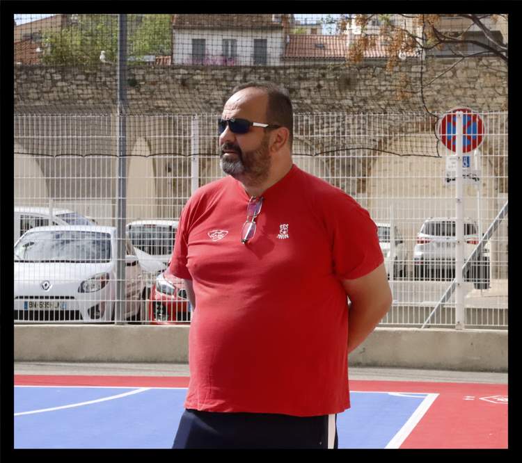 Un homme barbu portant des lunettes de soleil et un t-shirt rouge se tient sur un terrain de sport.