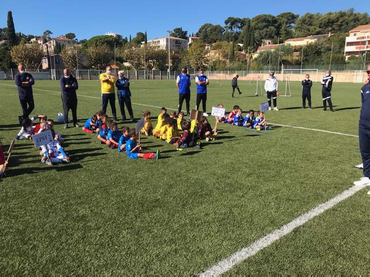 Des jeunes joueurs de football sont assis sur le terrain, encadrés par des adultes, sous un ciel bleu.