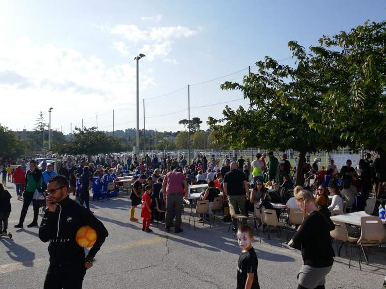 Une foule de personnes, dont des enfants en tenue de football, se rassemble autour de tables en extérieur, sous un ciel clair.