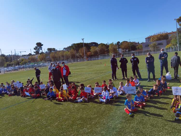 Des jeunes joueurs de football sont assis sur le terrain avec des adultes debout derrière eux, sous un ciel bleu.
