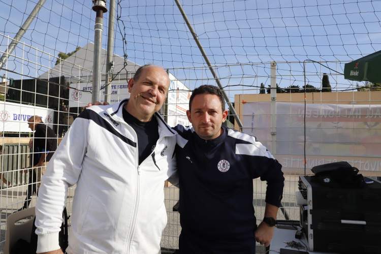 Deux hommes souriants posent côte à côte devant un filet de but et des installations de stade.