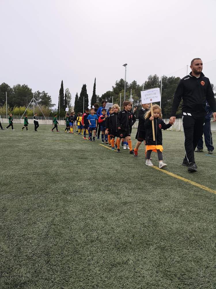 Des jeunes joueurs de football et leurs encadrants défilent sur un terrain en herbe synthétique.