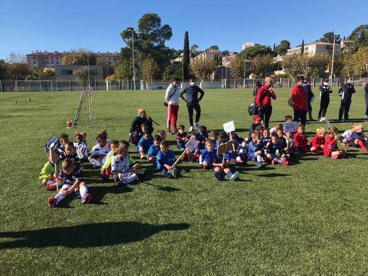 De jeunes footballeurs sont assis sur le terrain en herbe, entourés d'adultes, sous un ciel bleu.