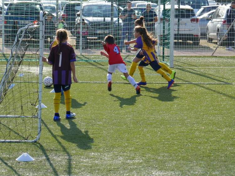 Des jeunes joueurs de football en pleine action sur un terrain synthétique, avec des voitures en arrière-plan.
