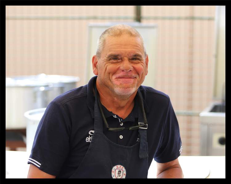 Un homme souriant porte un tablier et des lunettes autour du cou, dans un environnement de cuisine ou de buvette.