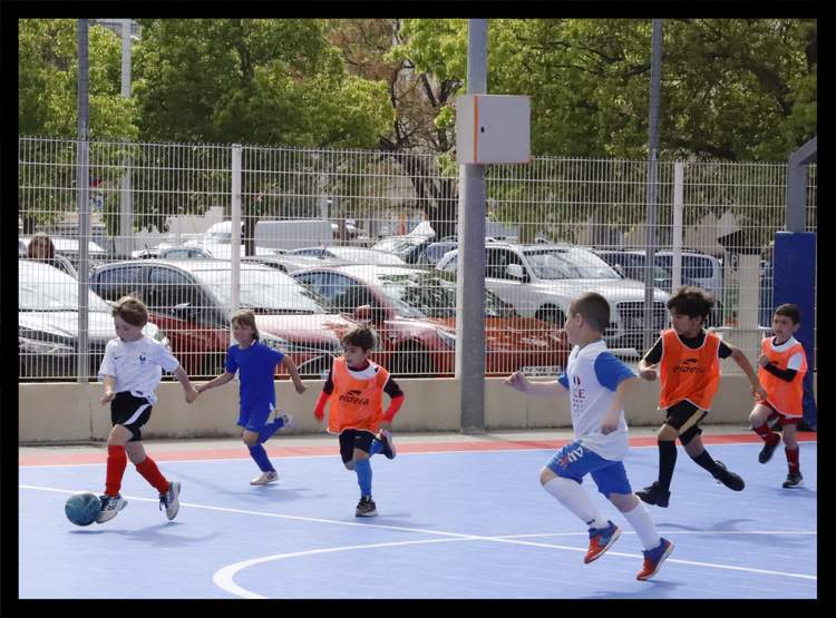 Des jeunes joueurs de football en pleine action sur un terrain bleu, avec un parking en arrière-plan.