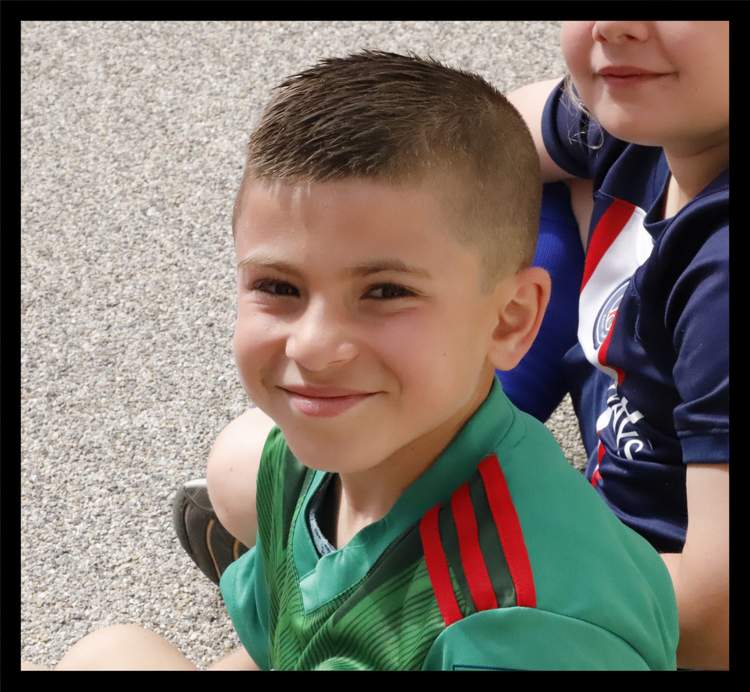 Un jeune garçon souriant en maillot de football vert et rouge, avec une autre personne partiellement visible à ses côtés.