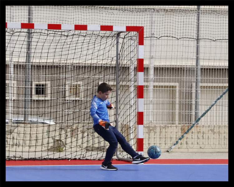 Un jeune gardien de but en maillot bleu s'apprête à frapper un ballon de football en salle devant un but.