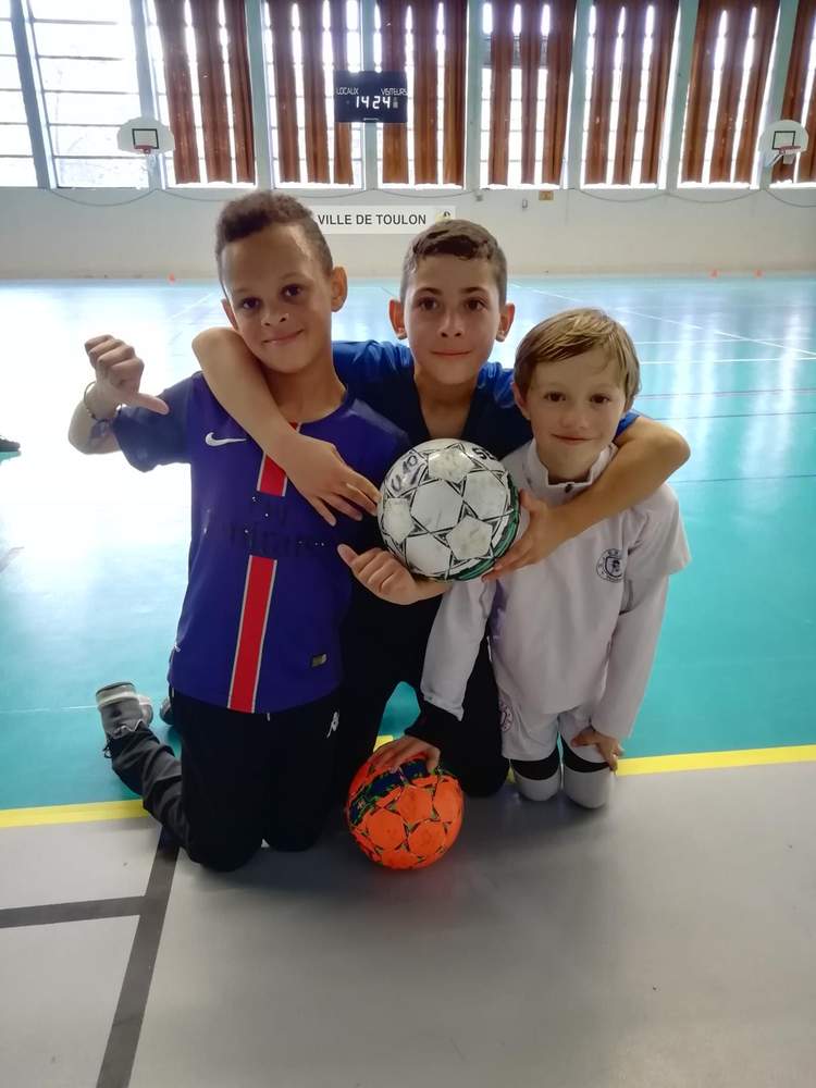 Trois jeunes joueurs de football posent ensemble avec des ballons dans un gymnase.