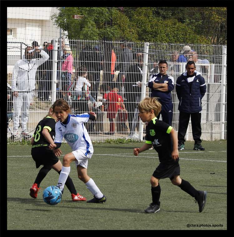 Des jeunes joueurs de football en pleine action sur le terrain, avec des adultes observant en arrière-plan.