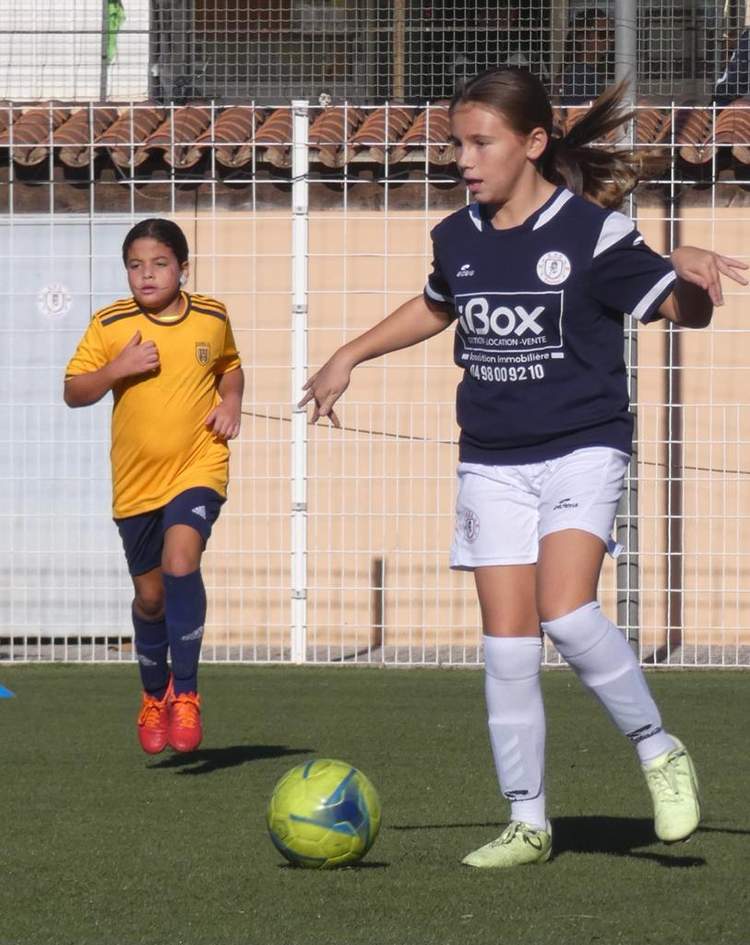 Deux jeunes joueuses de football en action sur un terrain synthétique, avec un ballon au premier plan.