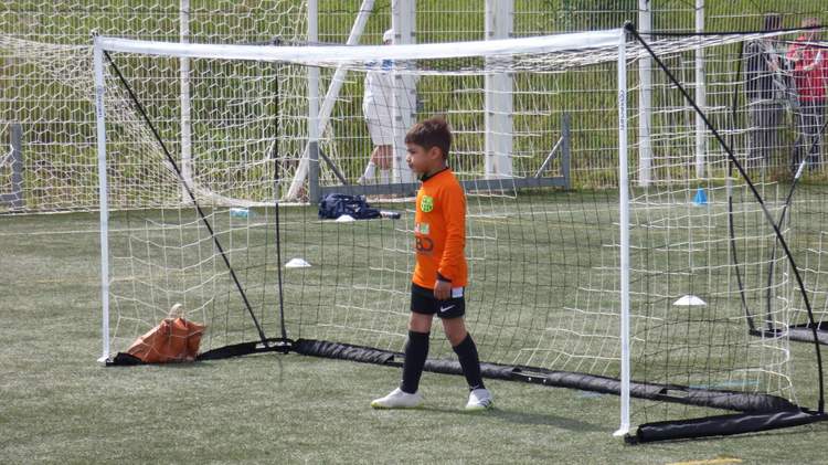 Un jeune joueur en maillot orange se tient devant un petit but de football sur un terrain synthétique.