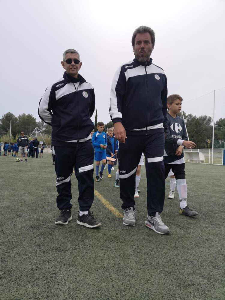 Deux entraîneurs adultes posent sur un terrain de football avec des jeunes joueurs en arrière-plan.