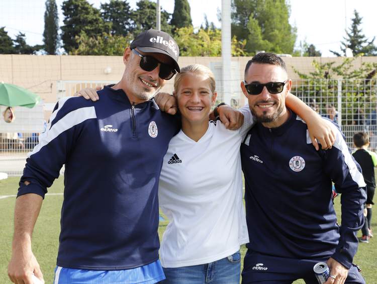 Trois personnes souriantes posent ensemble sur un terrain de football, deux hommes et une jeune femme.