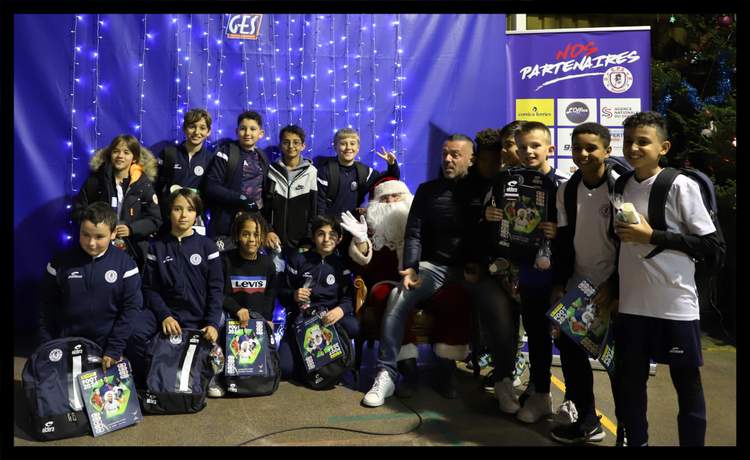 Un groupe de jeunes footballeurs pose avec le Père Noël et un adulte devant un mur décoré de lumières bleues et une bannière de partenaires.
