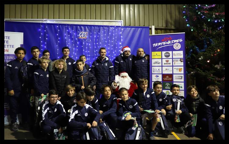Une équipe de jeunes footballeurs pose avec des adultes et le Père Noël devant un fond bleu et un sapin de Noël.