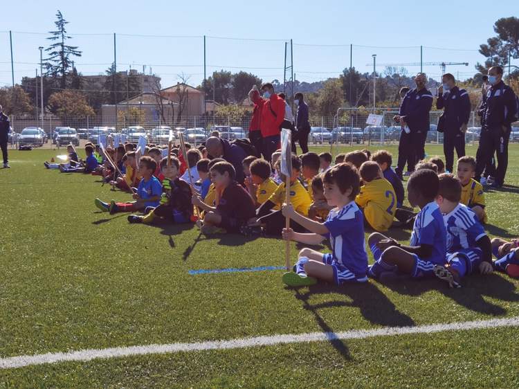 De jeunes footballeurs sont assis sur le terrain, écoutant attentivement des adultes debout en arrière-plan.