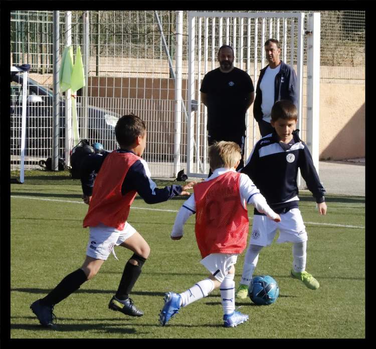 Des jeunes joueurs de football en pleine action sur un terrain, avec des adultes en arrière-plan.