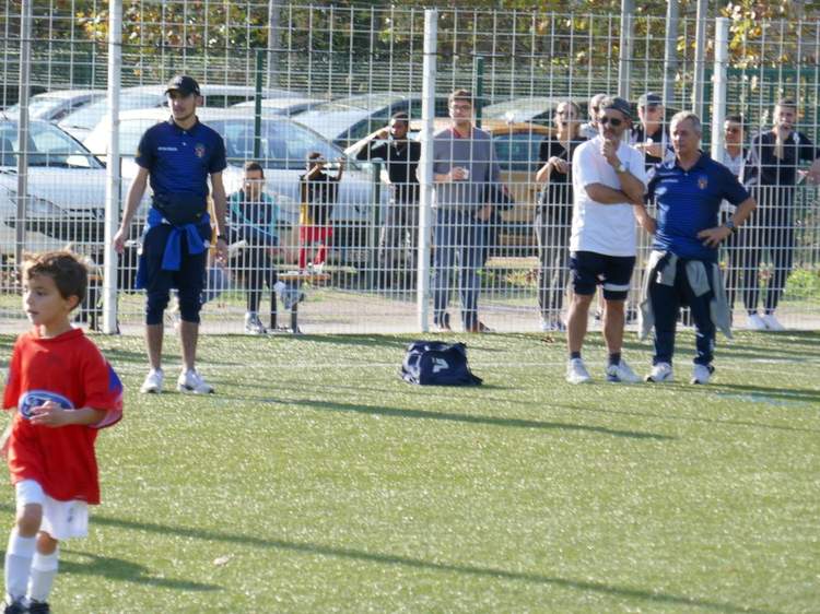 Un jeune joueur en maillot rouge au premier plan, avec des adultes et d'autres enfants derrière une clôture sur un terrain de football.