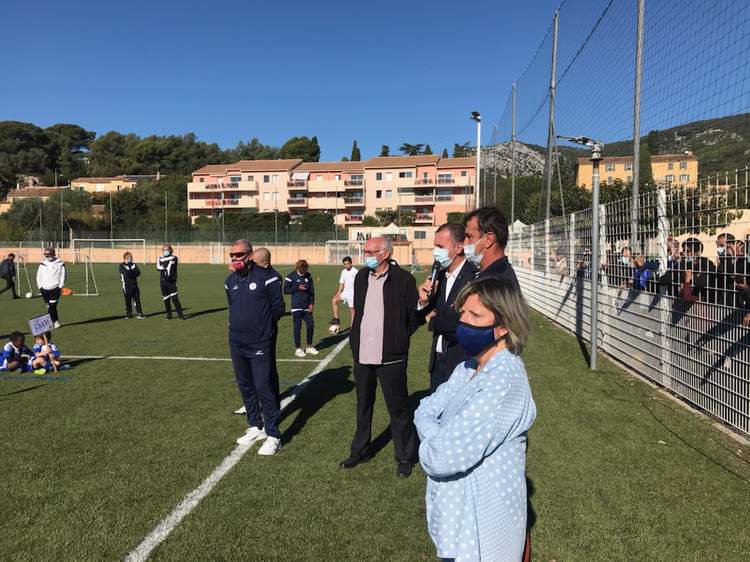 Des adultes et des enfants masqués se rassemblent sur un terrain de football en gazon synthétique sous un ciel bleu.