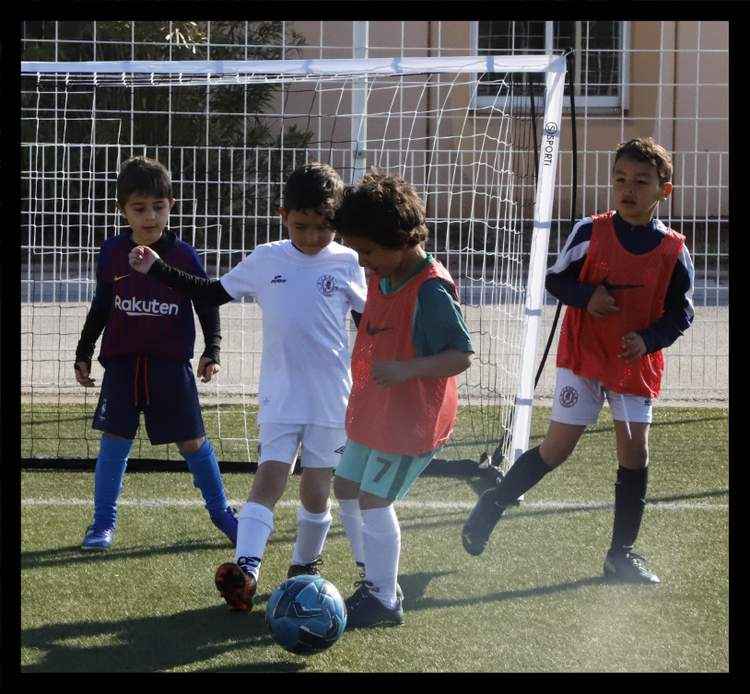 Quatre jeunes joueurs de football s'entraînent avec un ballon devant un but sur un terrain synthétique.
