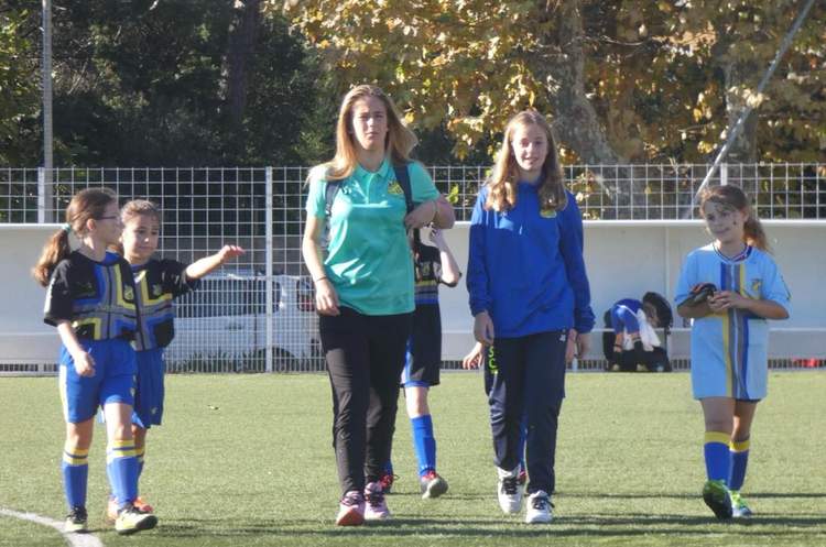 Des jeunes filles et des adultes marchent sur un terrain de football en gazon synthétique sous un soleil éclatant.