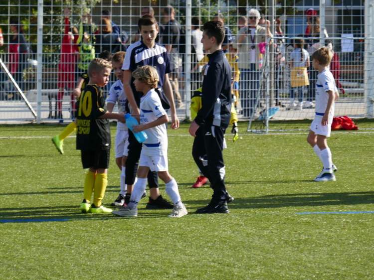 Des jeunes joueurs de football sur un terrain synthétique, certains avec des adultes en arrière-plan.