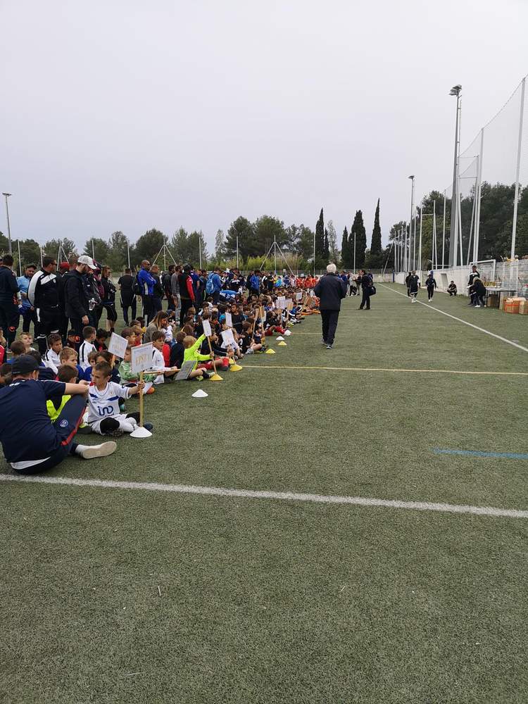 De nombreux jeunes joueurs de football sont assis sur le terrain, écoutant un adulte, lors d'un événement sportif.