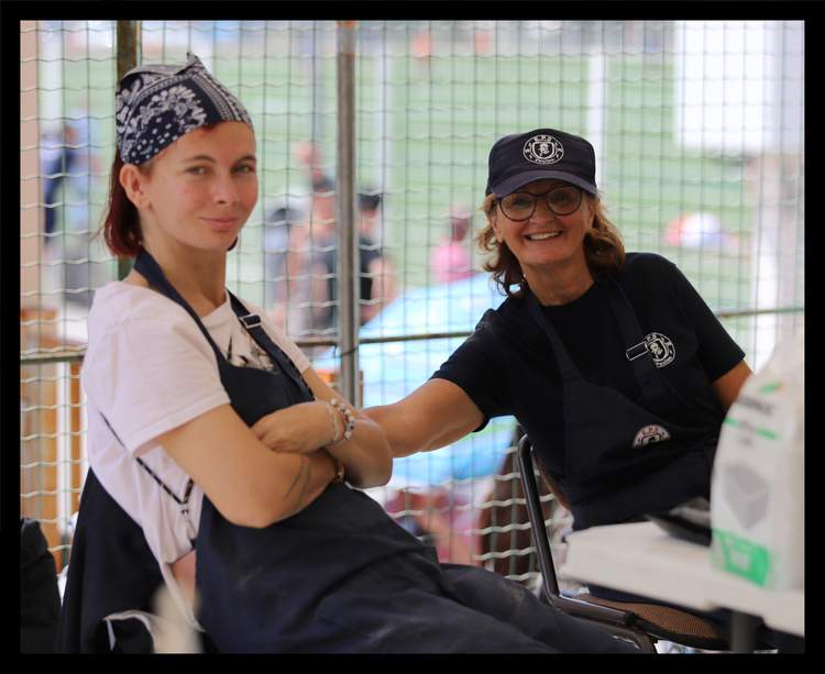 Deux femmes souriantes portent des tabliers et des casquettes, derrière une grille.