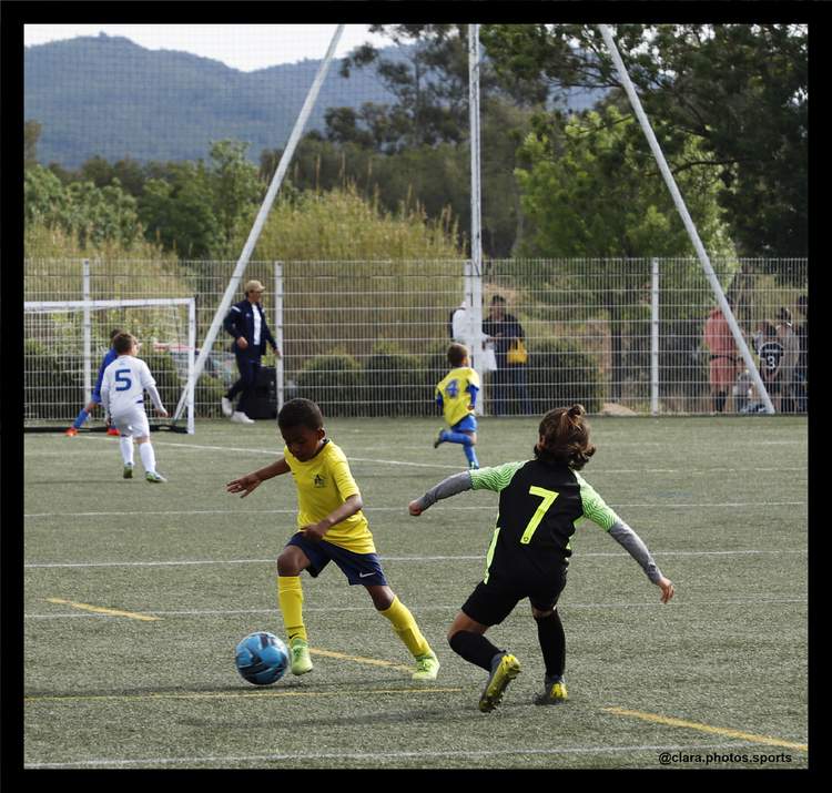 Des jeunes joueurs de football en pleine action sur un terrain, avec d'autres personnes en arrière-plan.