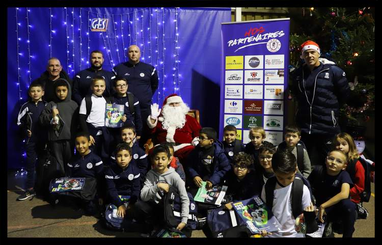 Des enfants et des adultes posent avec le Père Noël devant un mur décoré de lumières et une bannière de partenaires.